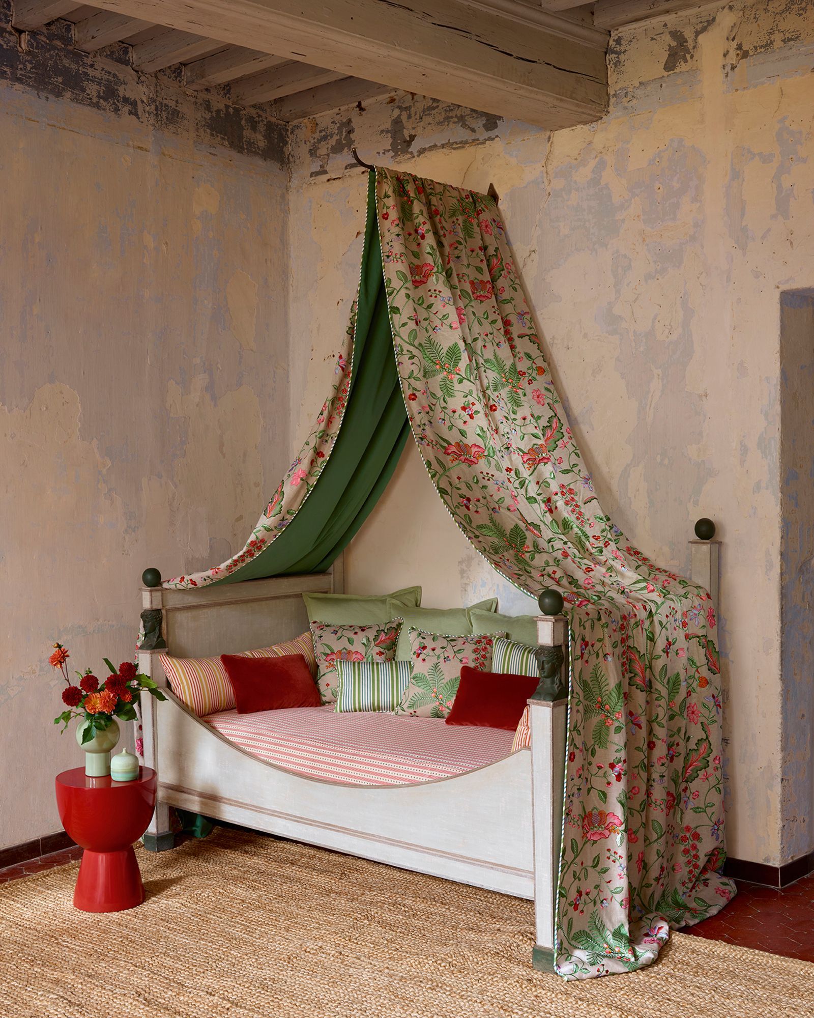 Bett mit grünem Baldachin und geblümtem Stoff, roten Akzenten und einem roten Beistelltisch auf einem Teppich.