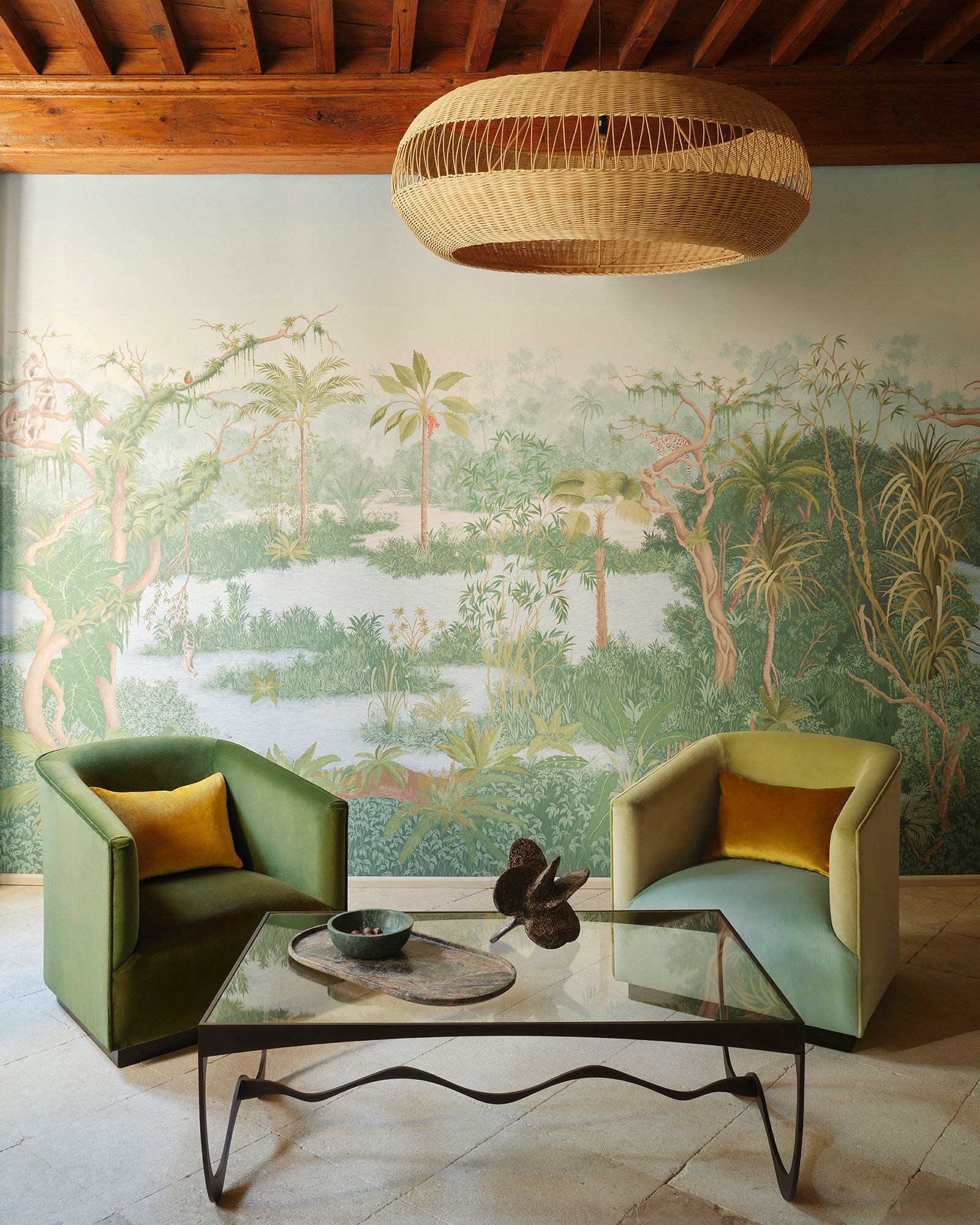Zwei grüne Sessel mit gelben Kissen stehen vor einem Couchtisch aus Glas, über dem eine geflochtene Lampe hängt. Eine malerische Tapete mit einer Landschaft im Hintergrund.