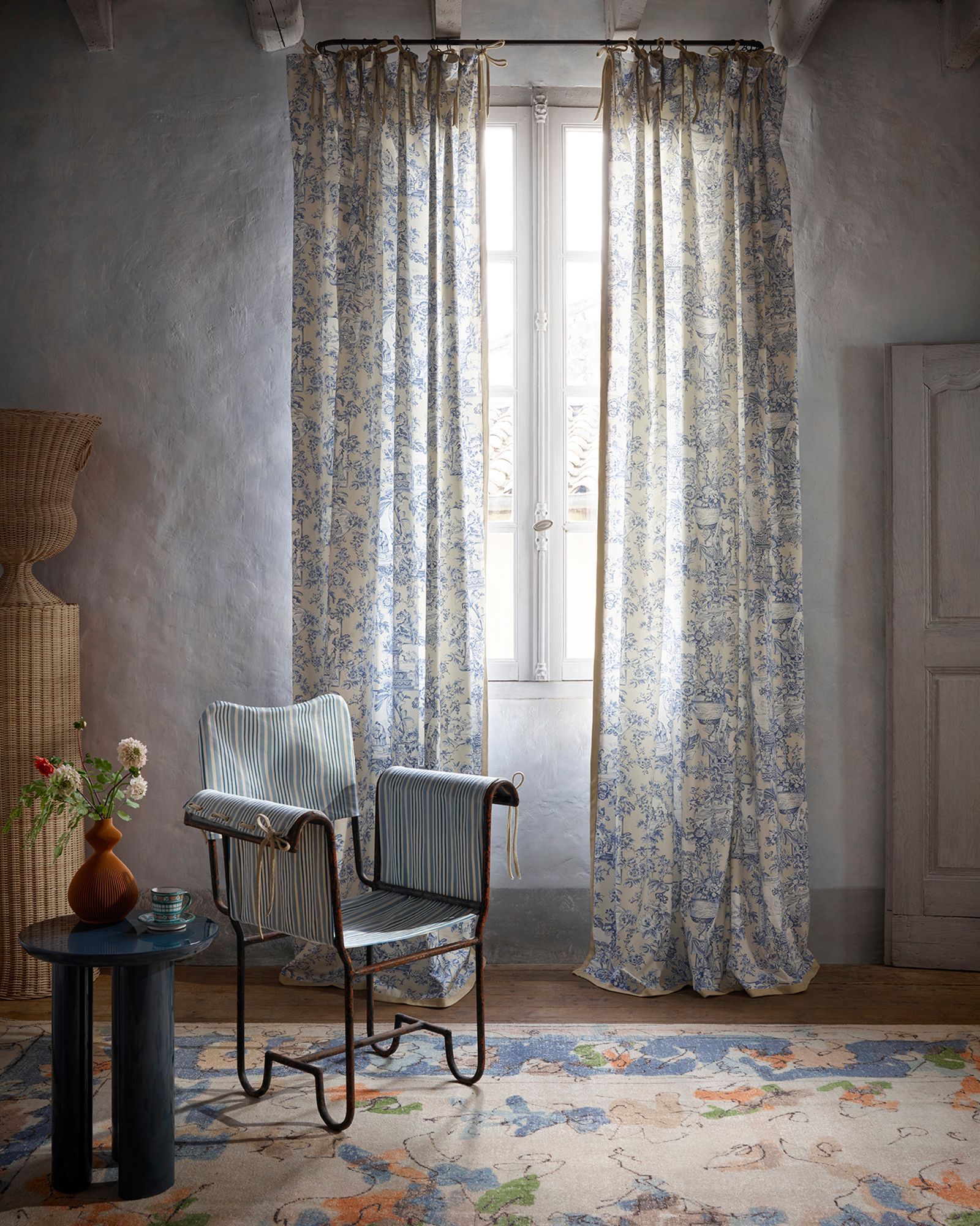 Ein Zimmer mit blauen Blumenvorhängen, Stuhl, Teppich und kleinem Tisch; Fenster mit weißem Rand.