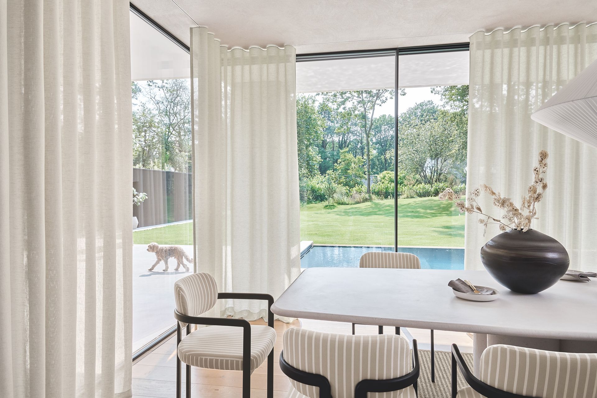 Esszimmer mit Tisch, Stühlen, Vorhängen und Blick auf einen Garten mit Pool; draußen läuft ein Hund.