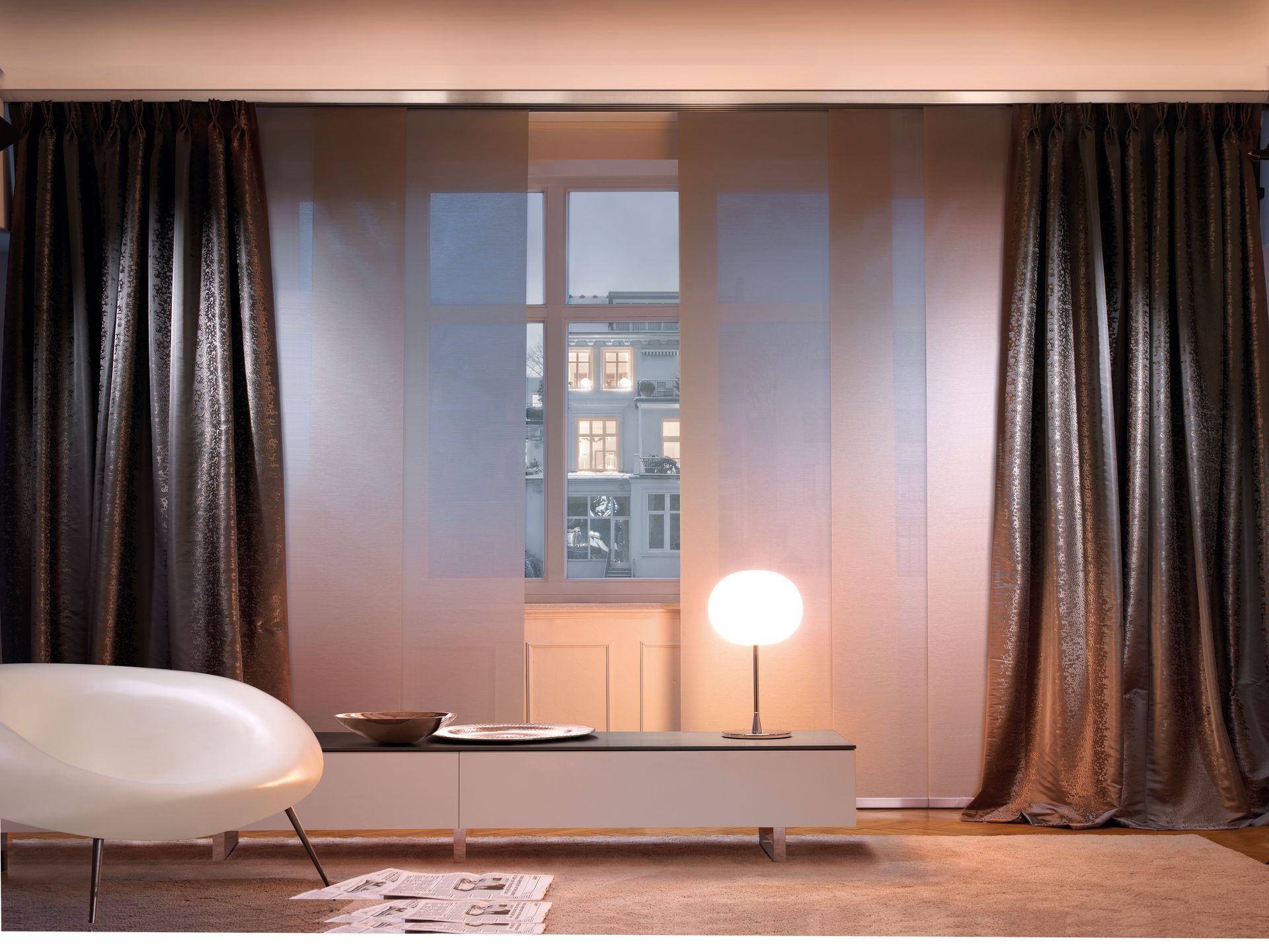 Wohnzimmer mit Fenster, transparenten Paneelen und dunkel gemusterten Vorhängen. Moderne Möbel, eine Lampe und Außengebäude sind sichtbar.