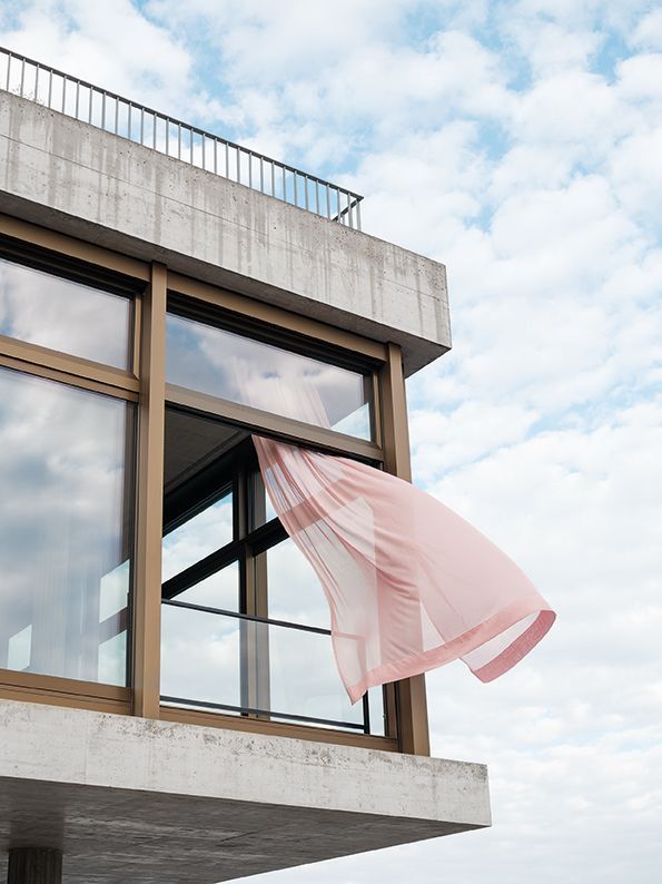 An einem teilweise bewölkten Tag weht ein rosa Vorhang aus einem offenen Fenster eines modernen Gebäudes.