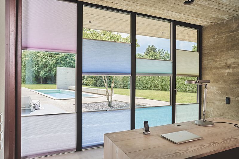Modernes Büro mit großem Fenster, das einen Pool zeigt. Mehrfarbige Jalousien verdecken das Fenster teilweise. Schreibtisch mit Laptop.