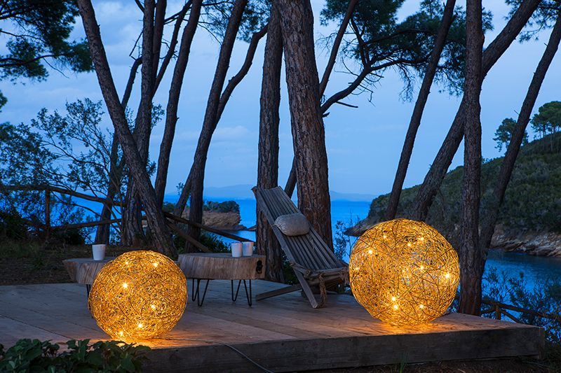 Zwei beleuchtete Kugellichter auf einem Deck mit Blick auf das Meer, Bäume und einen Liegestuhl in der Abenddämmerung.