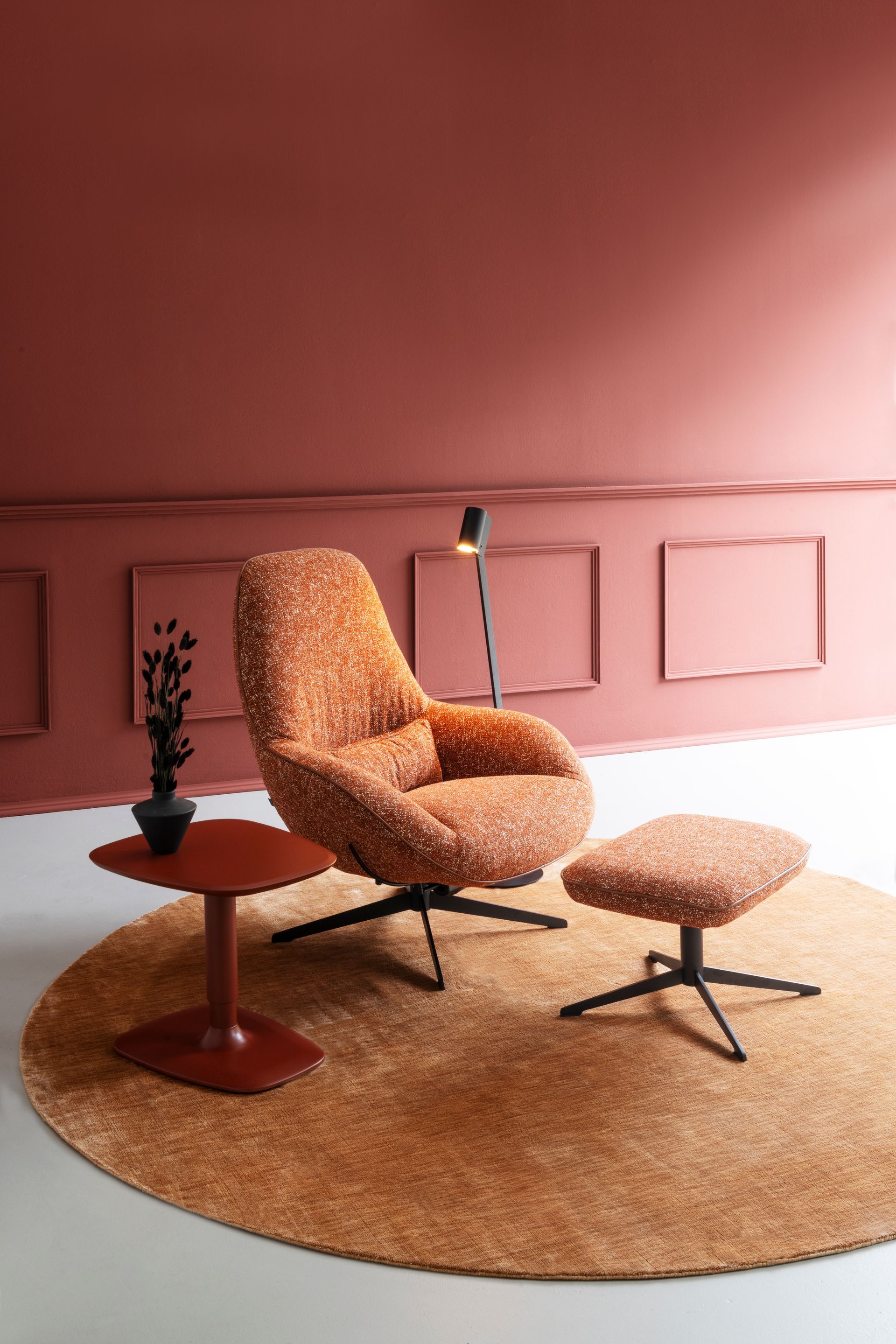 Gemütlicher Wohnraum mit einem orange gemusterten Sessel, einem Ottomanen und einem Teppich vor einer Terrakotta-Wand. Eine schwarze Lampe und ein Beistelltisch sind vorhanden.