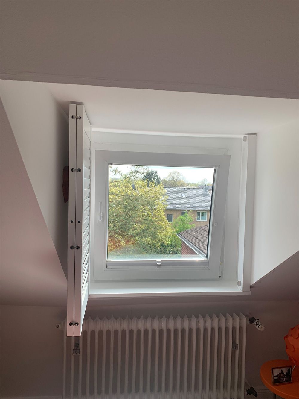 Weißes Dachfenster mit offenem Fensterladen, mit Blick auf einen baumbestandenen Hof und einem Heizkörper darunter.