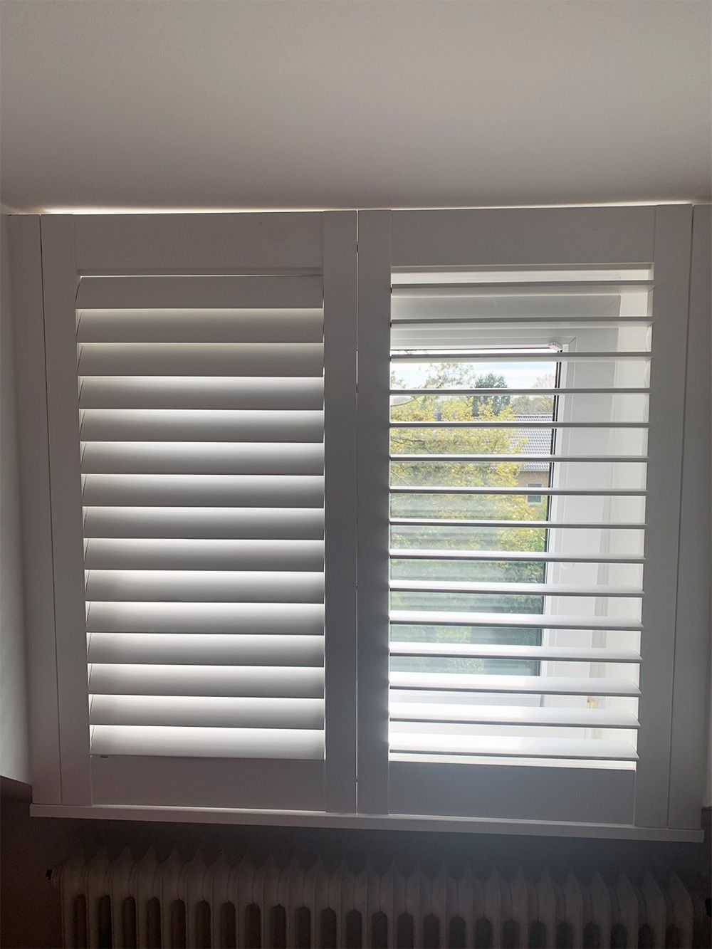 Weiße Fensterläden, eine Seite geschlossen, die andere offen, Blick auf Bäume möglich.