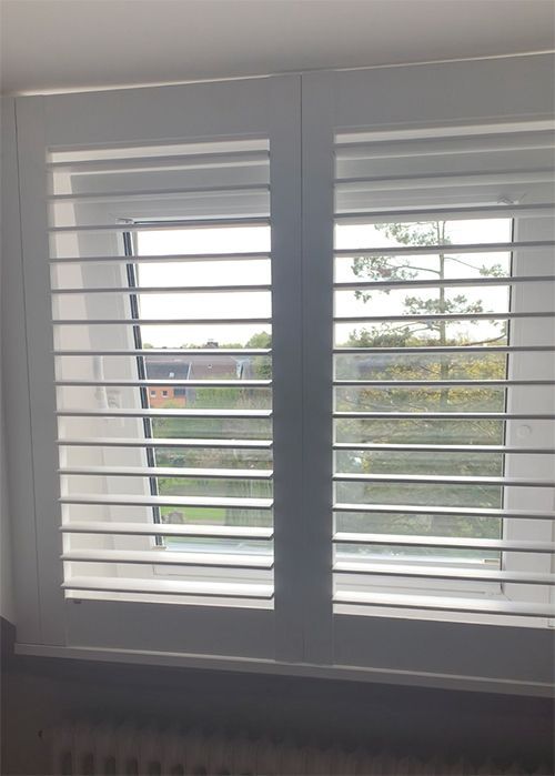 Weiße Fensterläden mit offenen Lamellen, die den Blick auf Bäume und ein Gebäude einrahmen.