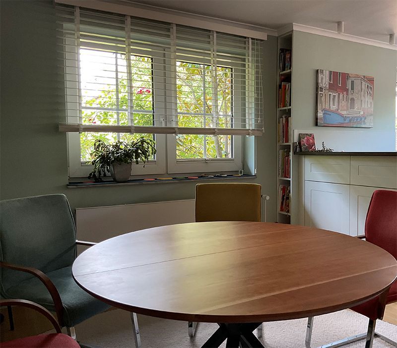 Runder Holztisch mit Stühlen vor einem Fenster mit Jalousien und einem Gemälde an der Wand.