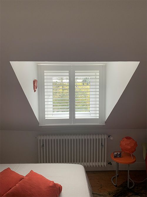 Ein Fenster mit weißen Fensterläden in einem dreieckigen Raum über einem Bett und einem orangefarbenen Stuhl in einem Zimmer.