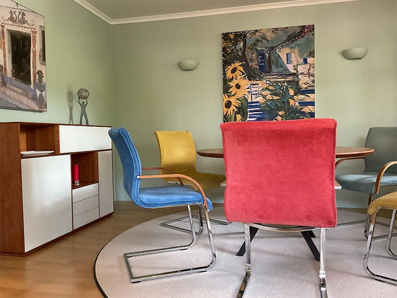 Esszimmer mit rundem Tisch, bunten Stühlen, Kunst und einem hölzernen und weißen Schrank.