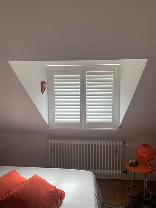 Schlafzimmer mit schrägem Fenster und weißen Fensterläden, Heizkörper, orangefarbenem Stuhl und Bett mit orangefarbenen Kissen.