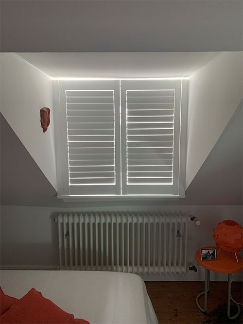 Weiße Fensterläden an einem Fenster in einem Raum mit Heizkörper, orangefarbenem Stuhl und roter Wanddekoration.