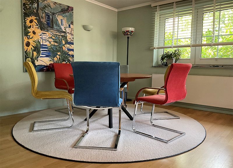 Esszimmer mit einem runden Tisch, vier bunten Stühlen auf einem runden Teppich. Kunstwerk und Fenster sichtbar.