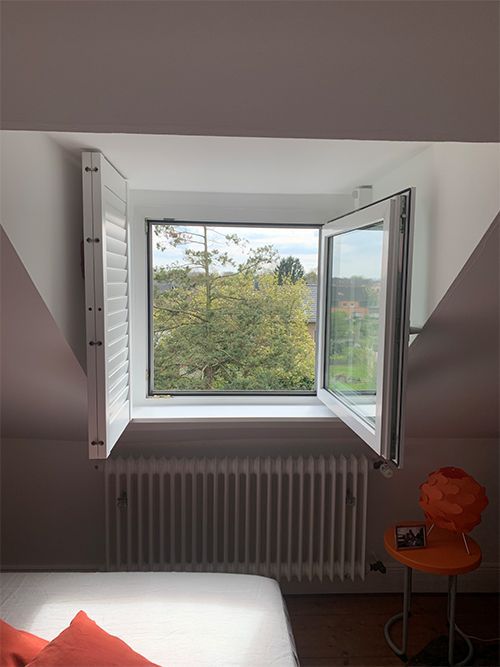 Fenster mit offenem Fensterladen, Blick ins Grüne. Weiße Wände und Heizkörper, orangefarbener Stuhl.