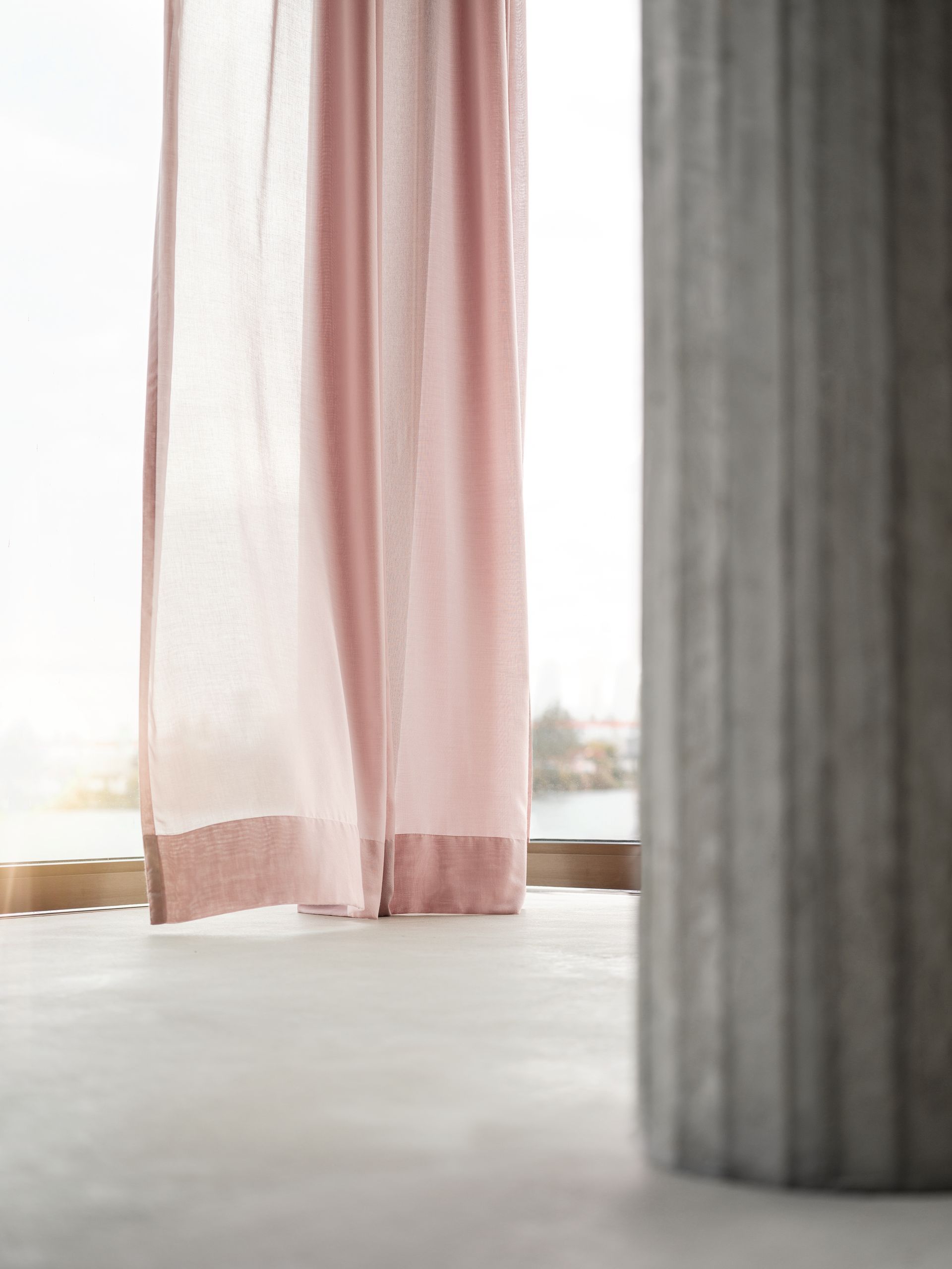 Ein rosafarbener, transparenter Vorhang hängt an einem Fenster, durch das Sonnenlicht fällt. Auf der rechten Seite befindet sich eine Betonsäule.