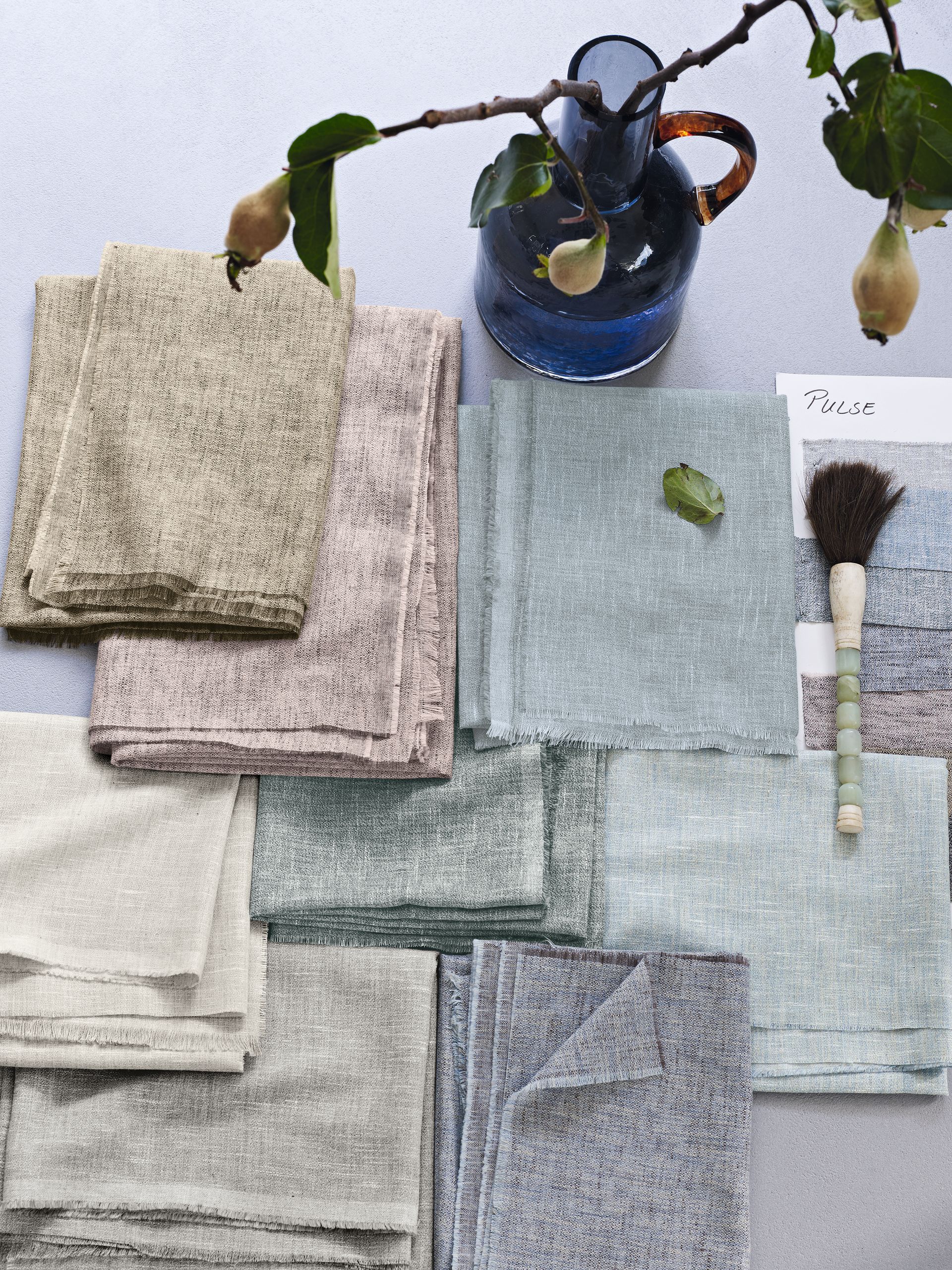 Gestapelte bunte Leinentücher mit einer blauen Vase, die einen Zweig hält.