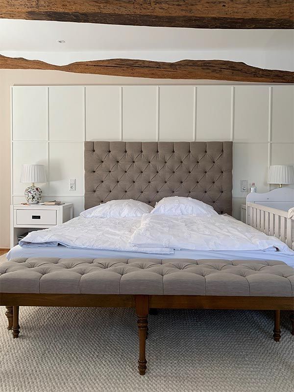 Schlafzimmer mit großem gepolstertem Kopfteil, weißer Bettwäsche, grauer Bank, Nachttischen und Kinderbett.