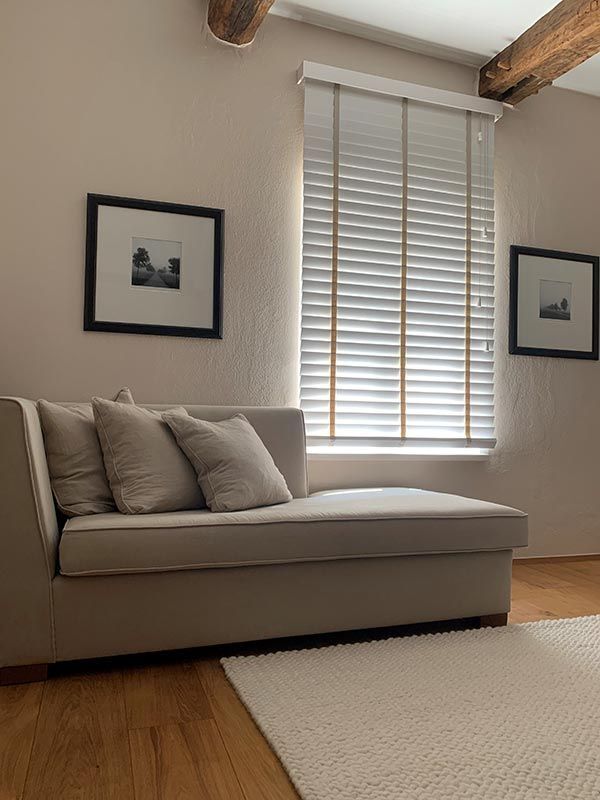 Beige Chaiselongue mit Kissen an einem Fenster mit Jalousien; zwei gerahmte Fotos hängen an einer hellen Wand.