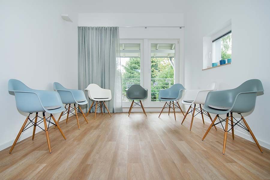 Kreis moderner Stühle in einem hellen Raum, nach außen gerichtet. Im Hintergrund sind ein Fenster und ein Vorhang zu sehen.