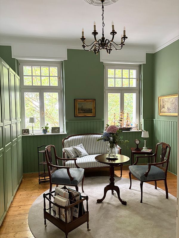 Elegantes grünes Wohnzimmer mit gestreiftem Sofa, Vintage-Stühlen, Kronleuchter und hohen Fenstern.