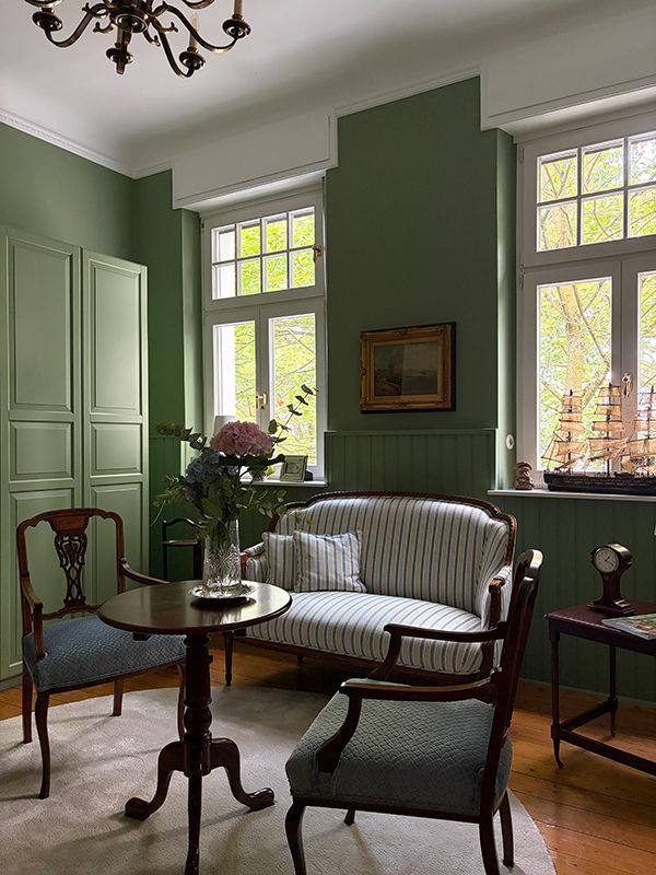 Elegantes grünes Wohnzimmer mit gestreiftem Sofa, Holzstühlen, kleinem Tisch und hohen Fenstern mit Tageslicht