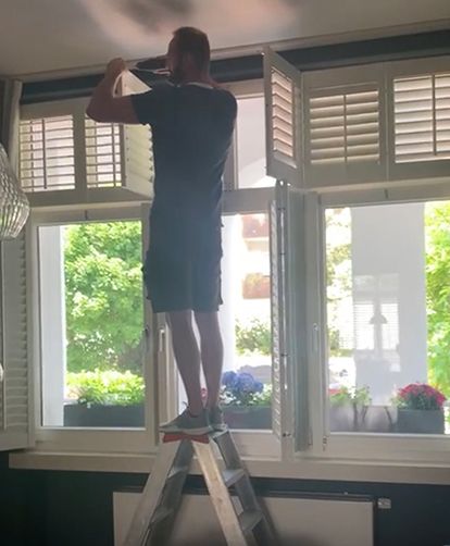 Mann auf einer Leiter, der Jalousien einstellt, weiße Fensterläden. Hell erleuchteter Raum, grüne Bäume draußen.