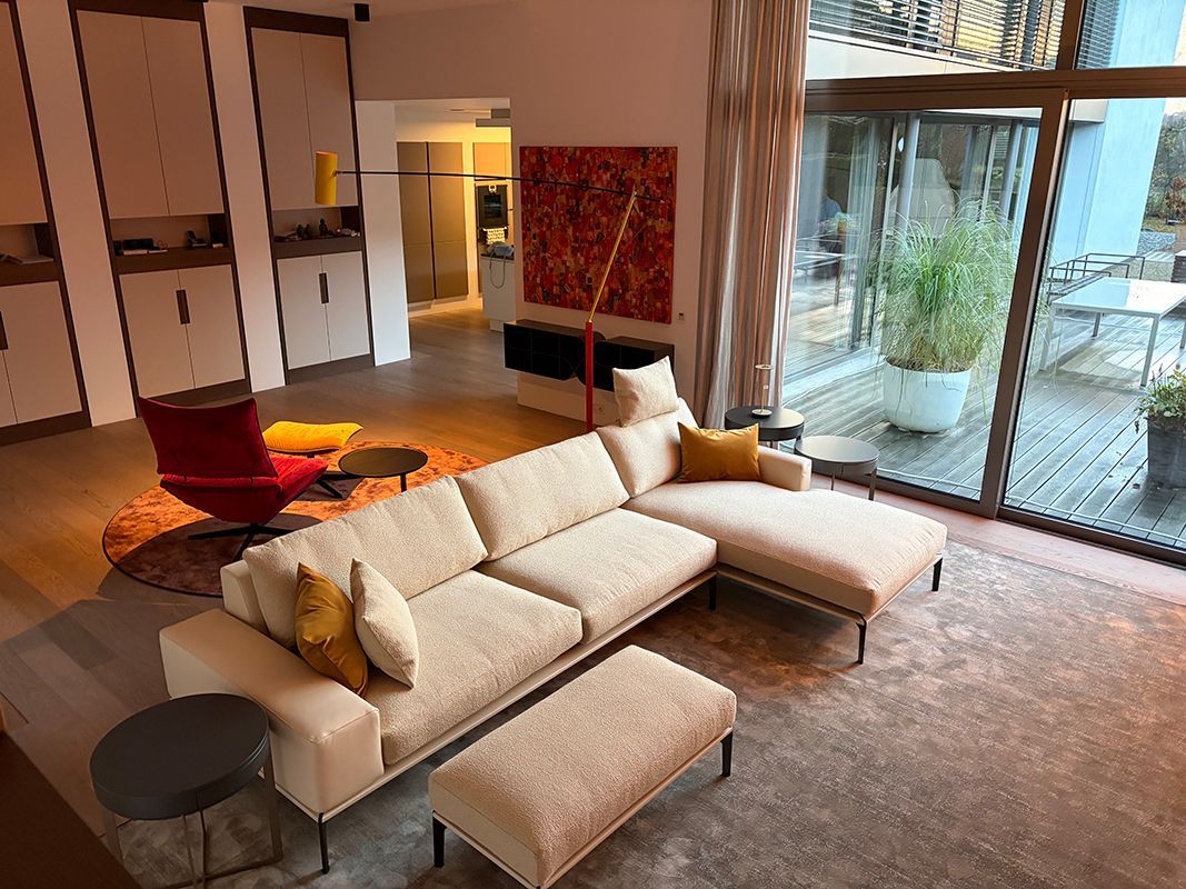 Wohnzimmer mit cremefarbenem Ecksofa, rotem Akzentstuhl und großen Fenstern mit Blick auf eine Terrasse.