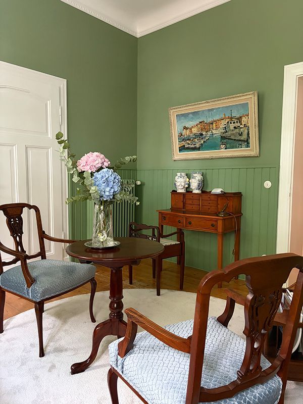 Grünes Wohnzimmer mit Holzstühlen, kleinem Tisch, Blumen und einem Landschaftsgemälde an der Wand.
