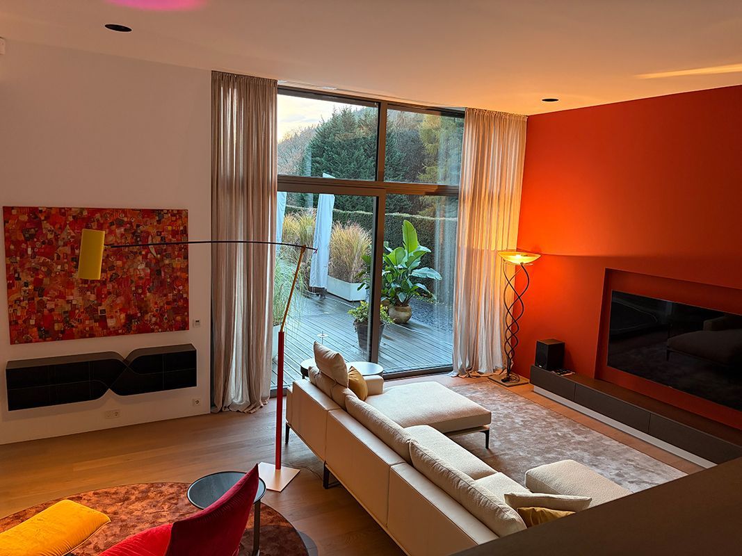 Modernes Wohnzimmer mit großem Fenster, weißem Sofa, orangefarbener Akzentwand und farbenfrohen Kunstwerken.