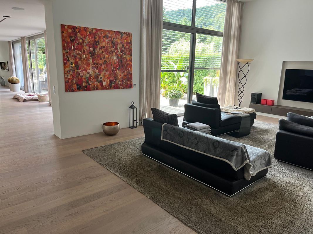 Wohnzimmer mit großen Fenstern und Blick auf einen Wald; schwarze Ledermöbel, ein großes abstraktes Gemälde und neutrale Farbtöne.