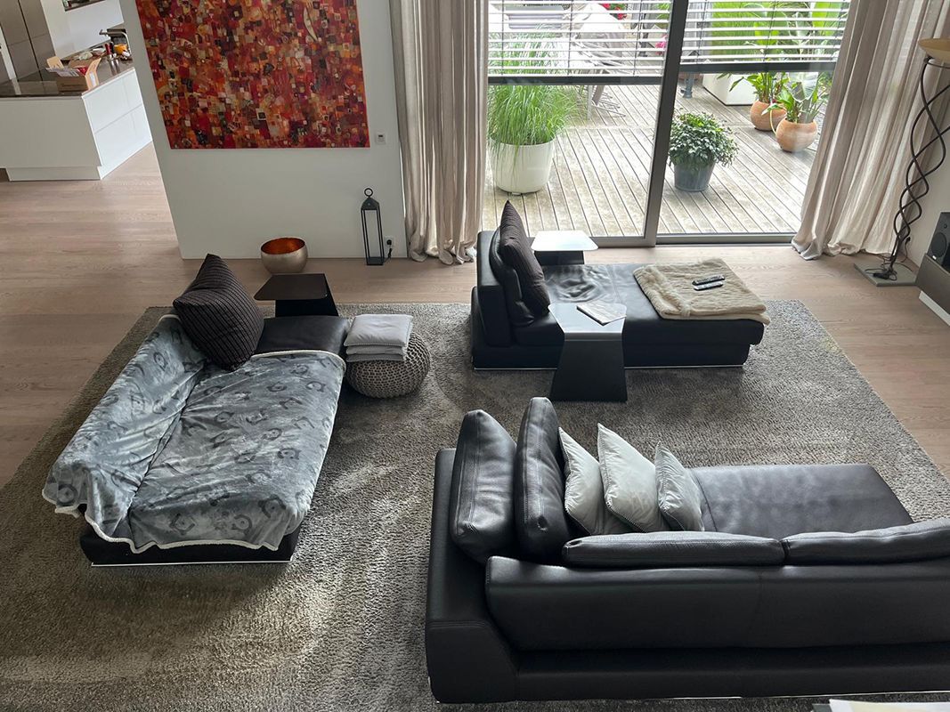 Wohnzimmer mit schwarzen Ledersofas auf einem grauen Teppich. Ein großes Fenster bietet Blick auf eine Terrasse mit Pflanzen.