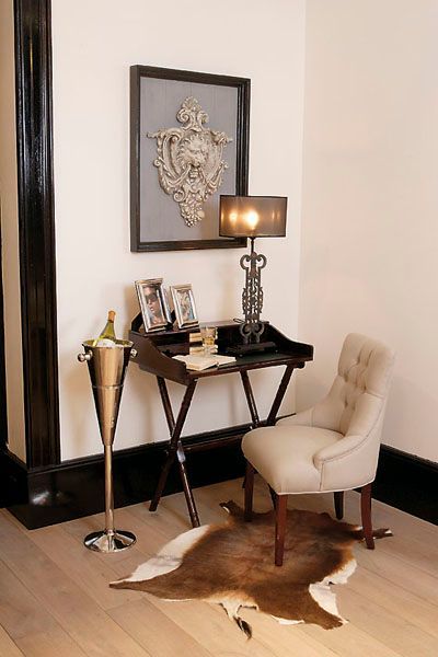 Eckvignette mit Schreibtisch, Stuhl, Kunst, Lampe, Wein und Kuhfellteppich. Schwarz, Weiß, Hellbraun.