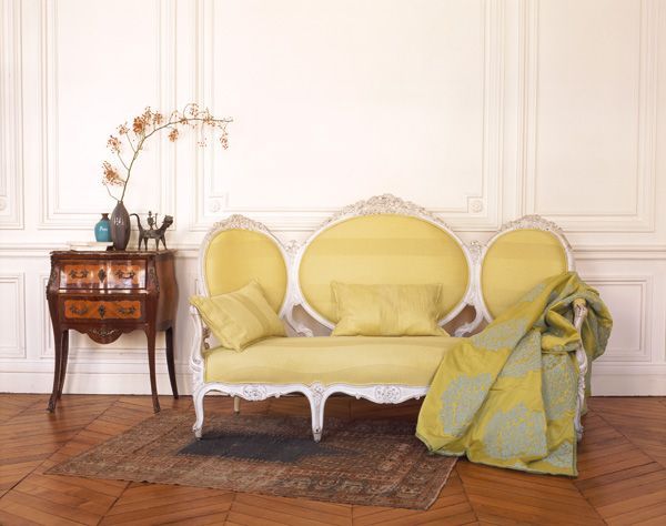 Gelbes Sofa vor einer weiß getäfelten Wand. Ein Beistelltisch mit Vase und Überwurfdecke auf einem Teppich.