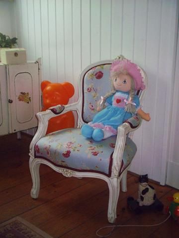 Verzierter Kinderstuhl mit geblümtem Stoff, auf dem eine Puppe sitzt. Orangefarbene Bärenfigur und Holzschrank im Hintergrund.