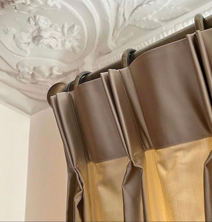 Nahaufnahme von taupefarbenen Vorhängen mit Falten, die an einer dunklen Stange vor einer weiß verzierten Decke hängen.