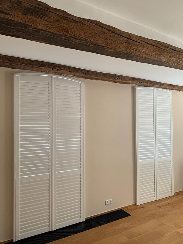 Weiße Lamellentüren vor einer hellen Wand mit Holzbalken darüber und Hartholzböden darunter.