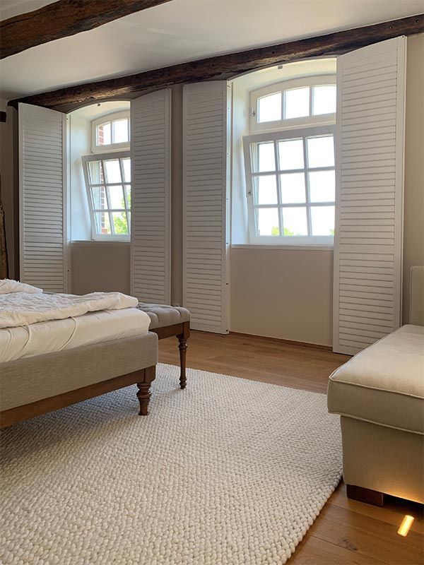 Schlafzimmer mit weißen Fensterläden, Holzbalken, beiger Bettwäsche und einem strukturierten weißen Teppich.