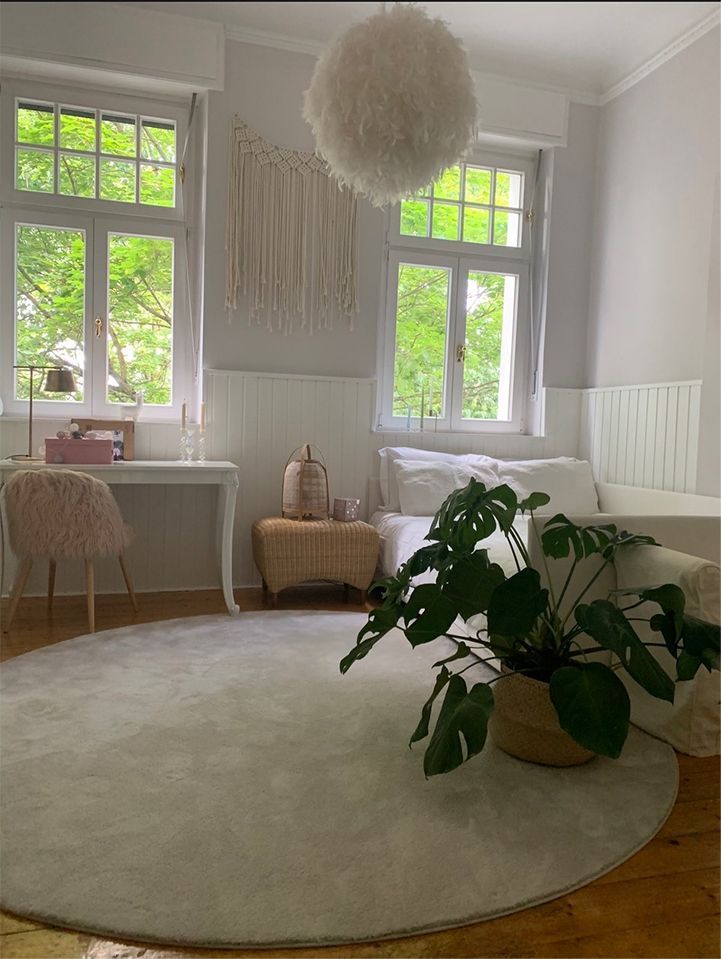 Helles, weißes Wohnzimmer mit großen Fenstern, rundem Teppich, üppiger Pflanze und gemütlicher Boho-Einrichtung