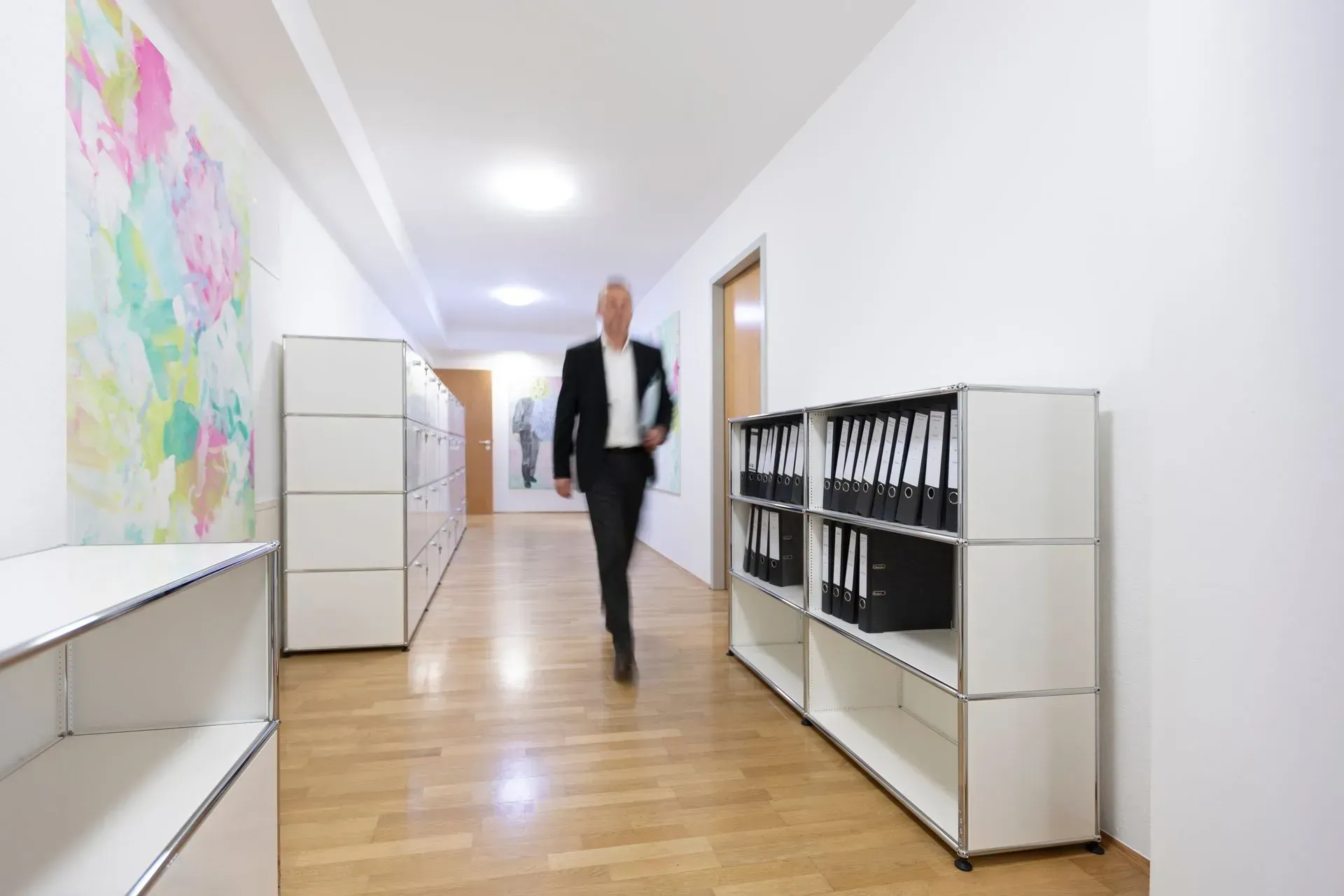 Eine Person geht einen hellen Büroflur mit weißen Schränken und Holzboden entlang.