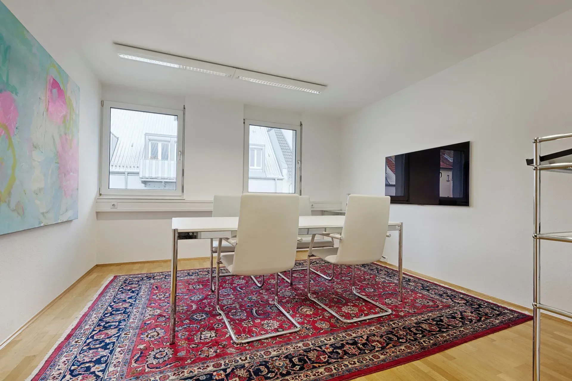 Helles, modernes Esszimmer mit Glastisch, weißen Stühlen, großem roten Teppich, Wandbildern und Fenstern.