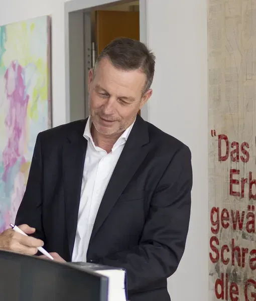 Ein Mann in einem dunklen Anzug unterzeichnet ein Dokument an einem Schalter in einem hellen Raum.
