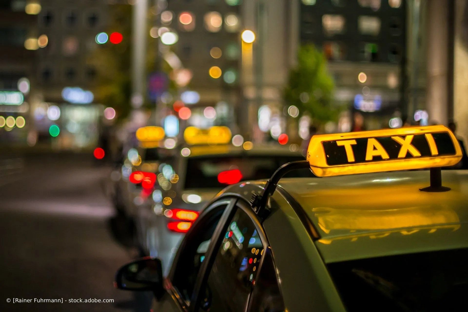 Eine Reihe gelber Taxis mit leuchtenden Dächern parkt nachts, vor verschwommenen Stadtlichtern.