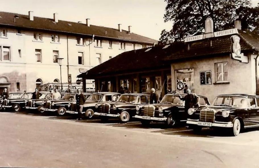 Eine Reihe dunkler Mercedes-Benz-Limousinen parkte vor einem Gebäude mit einem Schild; neben den Fahrzeugen standen Menschen.