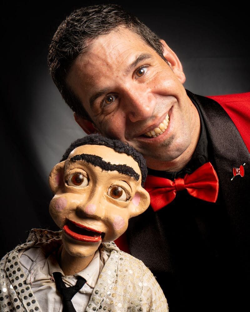 Un hombre con una chaqueta roja y pajarita sostiene una marioneta.