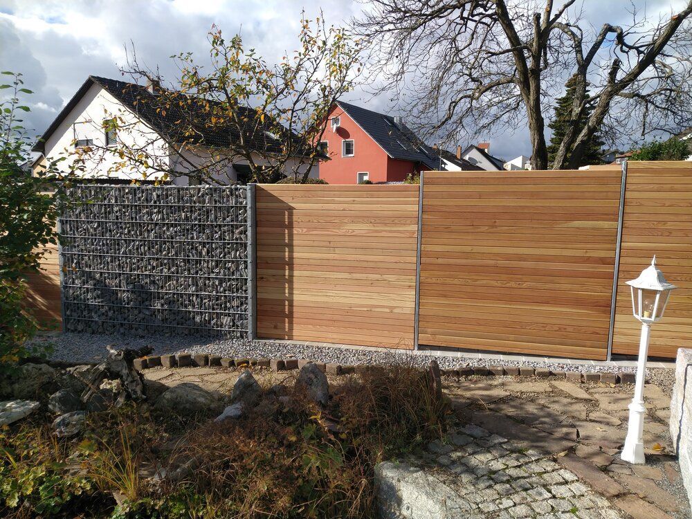 In einem Wohngarten steht ein Holzzaun mit einem mit Steinen gefüllten Metallgitterabschnitt. In der Nähe steht eine weiße Laterne.