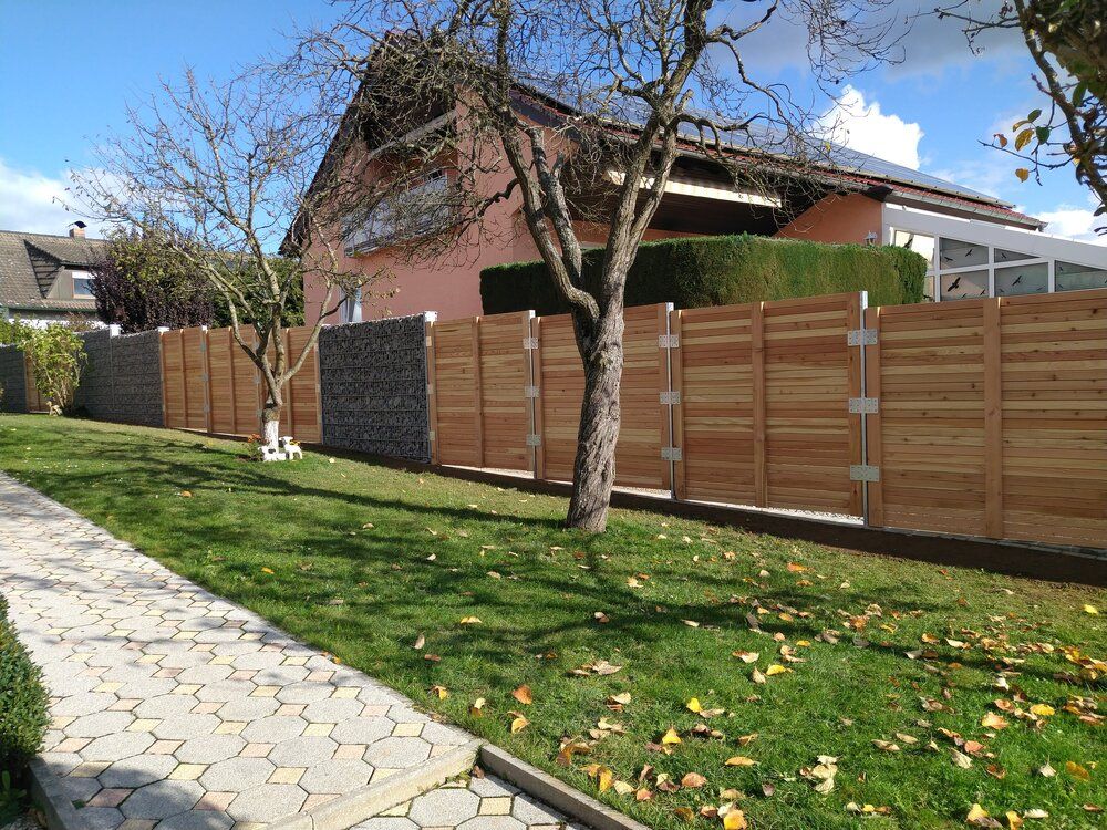 Ein Holzzaun umgibt ein Haus mit einem Steinabschnitt und einem Baum davor. Grüner Rasen, blauer Himmel.