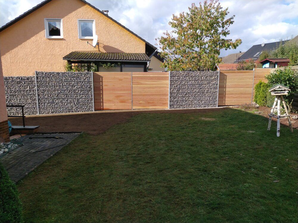 Ein Hinterhof mit einem Zaun aus Stein und Holz, grünem Rasen und einem beigen Haus im Hintergrund.