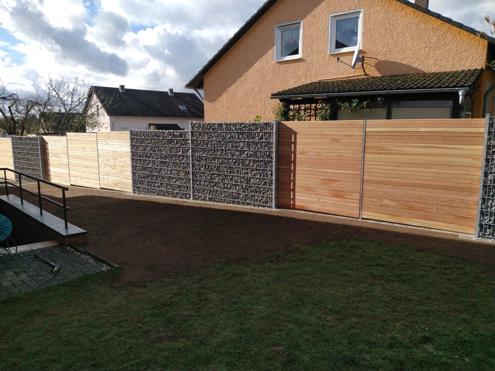 Hinterhof mit modernem Zaun: Holzpaneele und Gabionenelemente aus Stein, Rasenfläche, Haus im Hintergrund.