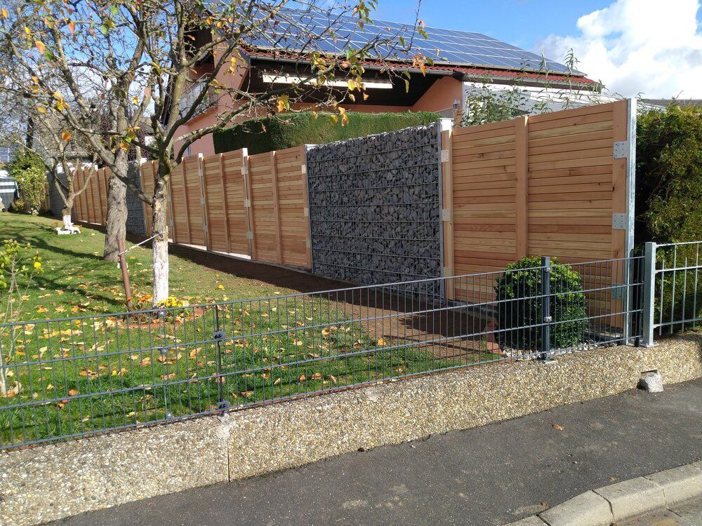 Ein Zaun aus Holz und Stein grenzt an einen grünen Rasen und eine gepflasterte Straße; im Hintergrund ist ein Haus zu sehen.